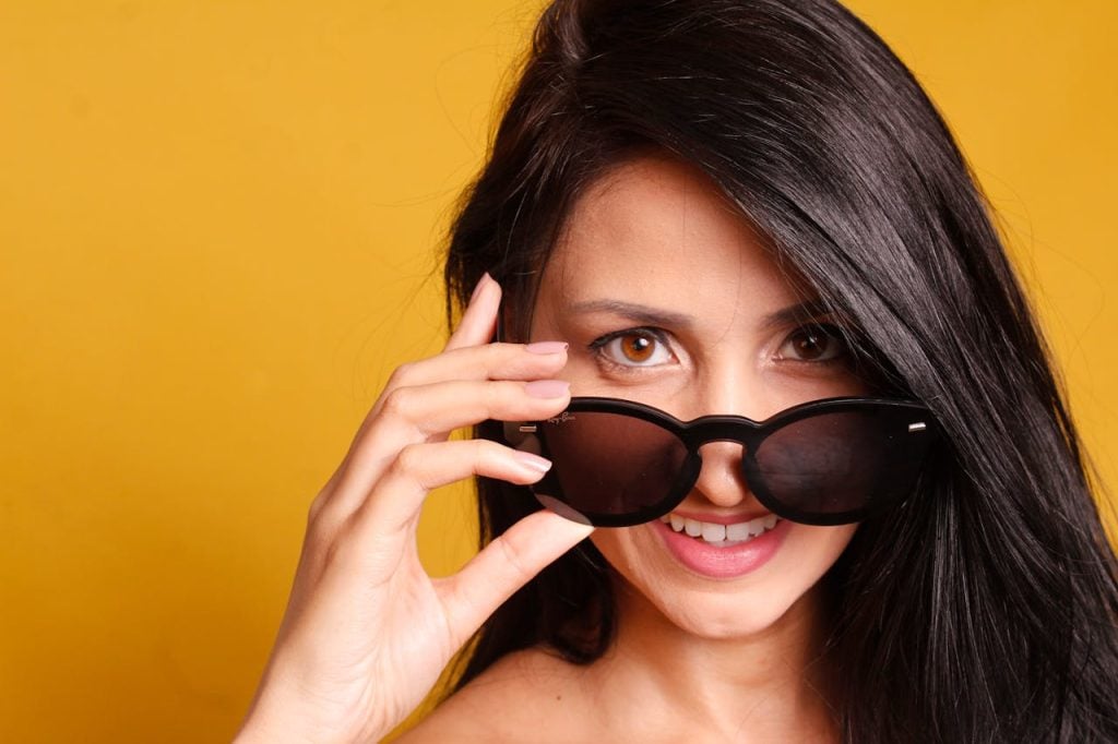 Portrait of a smiling woman with sunglasses posing against a vibrant yellow background, exuding style and confidence.