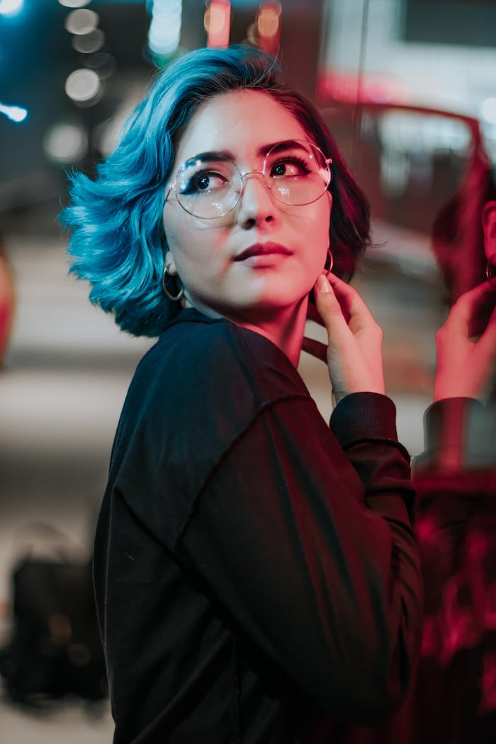 Vibrant portrait of an Asian woman with blue hair and glasses under colorful city lights.